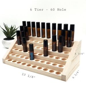 May include: A six-tier wooden essential oil display rack with 60 holes. The rack is 13 1/8 inches long, 6 1/4 inches high, and 4 3/4 inches deep. The rack holds several amber glass roller bottles with black caps. The text "6 Tier - 60 Hole" is visible.