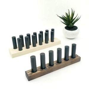Puede incluir: Dos porta-labiales de madera, uno de madera clara y otro de madera oscura. El porta-labiales de madera clara tiene 12 ranuras y el porta-labiales de madera oscura tiene 6 ranuras. Ambos porta-labiales están llenos de tubos de labial negro.