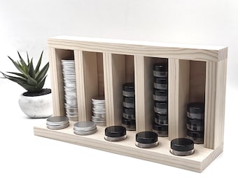 May include: A light-colored wooden storage rack with five vertical compartments. The rack holds stacks of small round containers with silver lids and stacks of small round containers with black lids, with additional containers placed on the base.