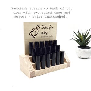 May include: A wooden display stand with two tiers, holding several black bottles. The stand has a light wood finish and a sign that reads "Superfine Pine, Fine Bottle Displays". A small potted succulent is to the right.