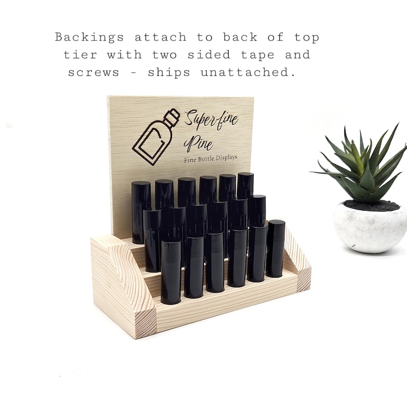 May include: A wooden display stand with two tiers, holding several black bottles. The stand has a light wood finish and a sign that reads "Superfine Pine, Fine Bottle Displays". A small potted succulent is to the right.