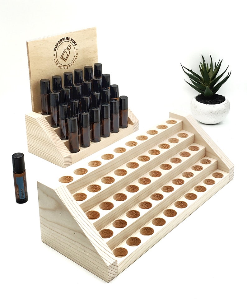 May include: Wooden essential oil display with multiple tiers. The display holds several small amber glass bottles with black caps. A small succulent plant in a white pot is in the background. The display has the text "SUPERFINE PINE FINE BOTTLE DISPLAYS".