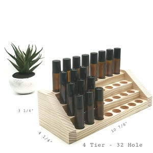May include: A wooden, 4-tier essential oil roller bottle display with 32 holes. The display holds several amber glass roller bottles with black caps. A small succulent plant in a white pot is to the left of the display.