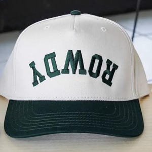 May include: A white baseball cap with a dark green brim and dark green embroidered text that reads ROWDY in an arched, upside-down orientation across the front panel. The cap features a structured crown with a top button and stitched detailing.