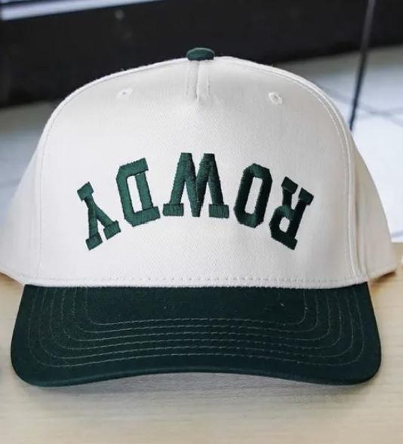 May include: A white baseball cap with a dark green brim and dark green embroidered text that reads ROWDY in an arched, upside-down orientation across the front panel. The cap features a structured crown with a top button and stitched detailing.