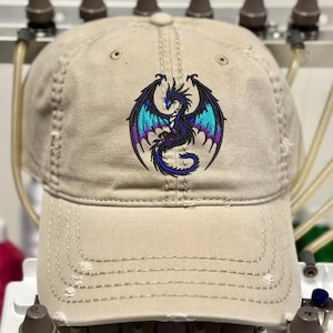 May include: Tan baseball cap with a distressed finish. The cap features an embroidered dragon design in black, teal, and purple. The dragon has outstretched wings and a curved tail. The cap has a curved brim and an adjustable strap.