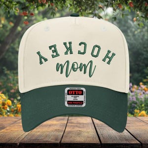 May include: A cream-colored baseball cap with a dark green brim. The front features the word HOCKEY in dark green block letters and the word mom in dark green cursive script. A rectangular sticker on the brim reads OTTO CLASSIC FIT OSFM.