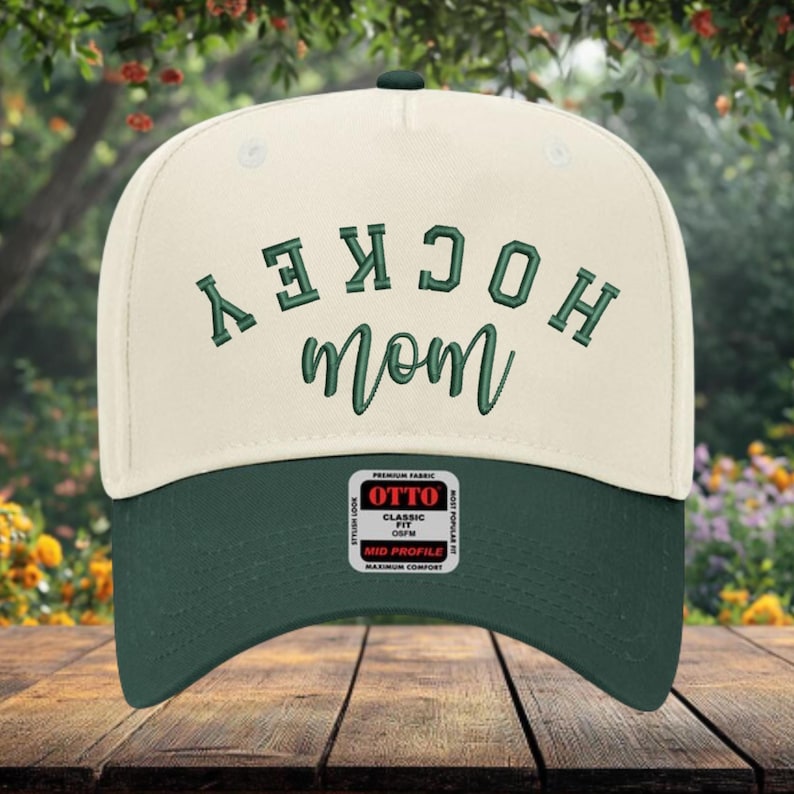 May include: A cream-colored baseball cap with a dark green brim. The front features the word HOCKEY in dark green block letters and the word mom in dark green cursive script. A rectangular sticker on the brim reads OTTO CLASSIC FIT OSFM.