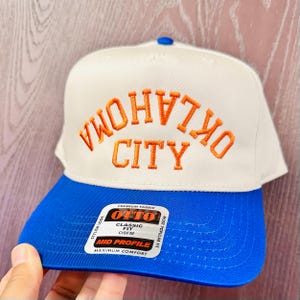 May include: A white and blue baseball cap with "OKLAHOMA CITY" embroidered in orange. The cap has a blue brim and a white crown. The cap has a "MID PROFILE" tag.