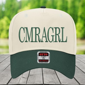 May include: A cream-coloured baseball cap with a dark green peak. The embroidered text "CMRAGRL" is in dark green. The cap has an "OTTO" tag with text that reads "Classic Fit OSFM Mid Profile Maximum Comfort".