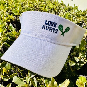 May include: White visor with a blue embroidered message that says "LOVE HURTS" and two green tennis rackets.