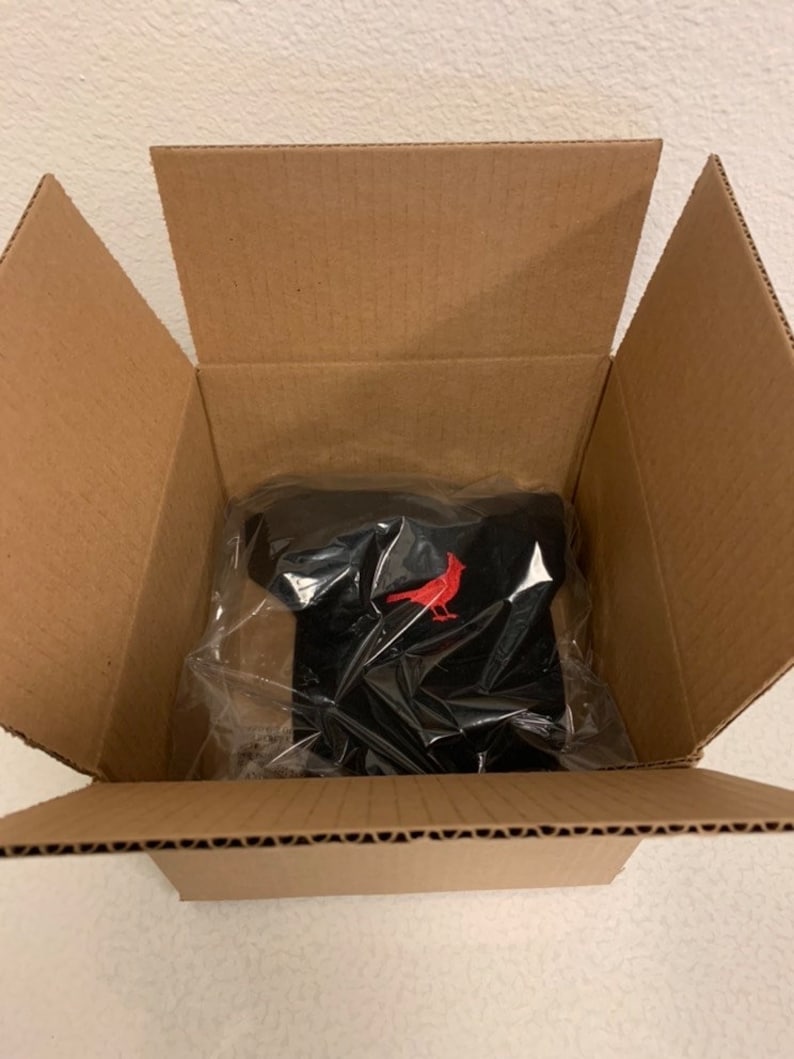 May include: A black t-shirt with a red cardinal logo, packaged in clear plastic inside a brown cardboard box. The box is open, revealing the shirt. The shirt is folded neatly.