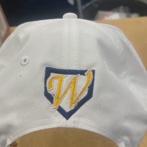 May include: A white baseball cap featuring an embroidered logo on the front. The logo consists of a yellow letter W outlined in white, centred inside a dark blue home plate shape. The cap includes a top button and ventilation eyelets.