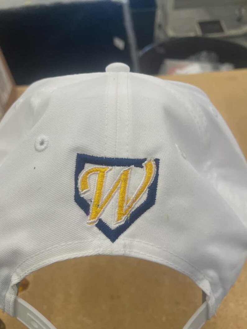 May include: A white baseball cap featuring an embroidered logo on the front. The logo consists of a yellow letter W outlined in white, centred inside a dark blue home plate shape. The cap includes a top button and ventilation eyelets.