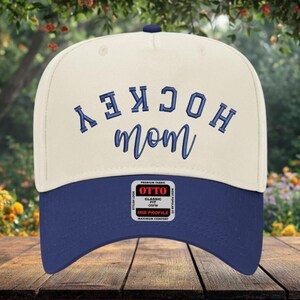 May include: A cream-colored baseball cap with a navy blue brim and a navy blue button on top. The front features the embroidered text HOCKEY in a block font and mom in a cursive script. A rectangular sticker on the brim reads OTTO CLASSIC FIT.