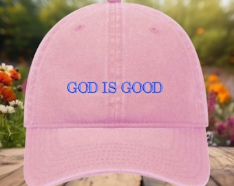 May include: A pale pink baseball cap with the blue text "GOD IS GOOD". The cap features a curved peak and a fabric strap at the back. The hat is made from a soft, textured material.