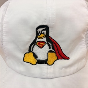 May include: A white baseball cap featuring an embroidered graphic of a penguin wearing a red cape and a chest emblem with a yellow letter S. The cap is constructed from a textured white fabric with mesh side panels and a structured front crown.
