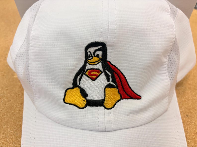 May include: A white baseball cap featuring an embroidered graphic of a penguin wearing a red cape and a chest emblem with a yellow letter S. The cap is constructed from a textured white fabric with mesh side panels and a structured front crown.