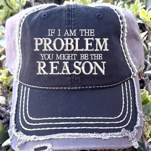 Peut inclure: Une casquette de baseball noire et grise usée avec le texte "If I am the problem you might be the reason" brodé en blanc.
