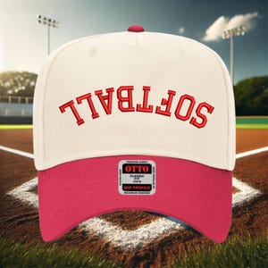 May include: A white and pink baseball cap with the word "SOFTBALL" embroidered in red on the front. The cap has a mid-profile and a classic fit.
