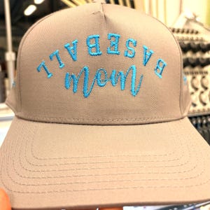 May include: A light brown baseball cap featuring inverted embroidery in bright blue thread. The text reads BASEBALL in a block font arched over the word mom in a cursive script. The embroidery is centered on the front panel of the cap.