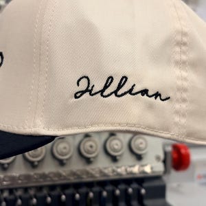 May include: A cream-colored baseball cap with a black brim. The side of the cap features the name Jillian embroidered in black cursive script. The cap is positioned in front of an embroidery machine with visible needles and mechanical components.