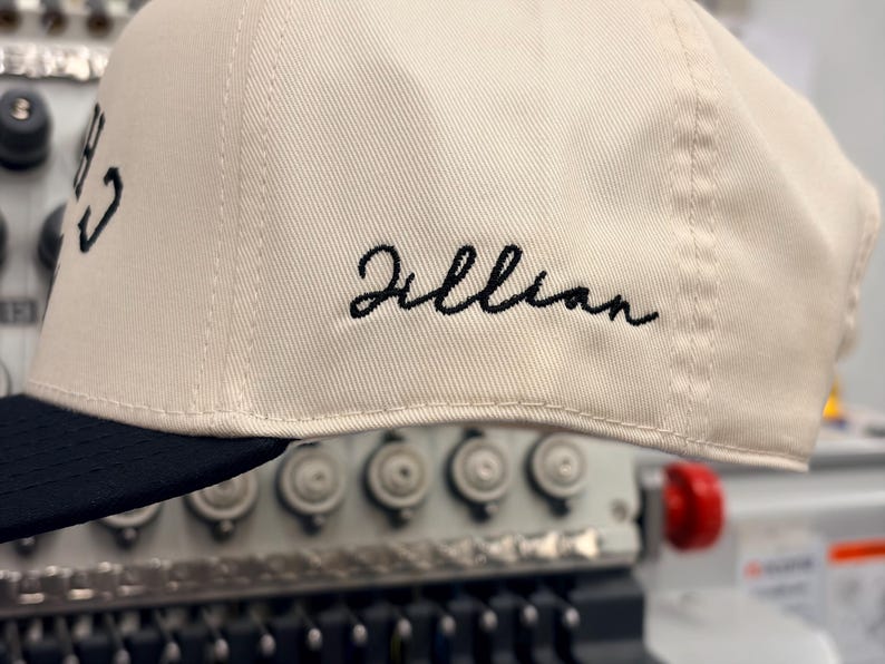 May include: A cream-colored baseball cap with a black brim. The side of the cap features the name Jillian embroidered in black cursive script. The cap is positioned in front of an embroidery machine with visible needles and mechanical components.