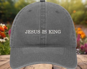 May include: A gray baseball cap with the text "JESUS IS KING" in white. The cap has a curved brim and a fabric strap closure. The background is a blurred view of flowers and foliage.