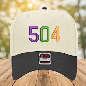 May include: A baseball cap with a cream-colored crown and a black brim. The cap features the number "504" embroidered in purple, green, and gold. The cap has a "OTTO" tag on the front.