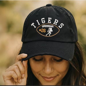 May include: A black distressed baseball cap featuring an embroidered design with the word TIGERS in white arched lettering above a brown football graphic containing the number 26 in yellow and a white silhouette of a football player running.