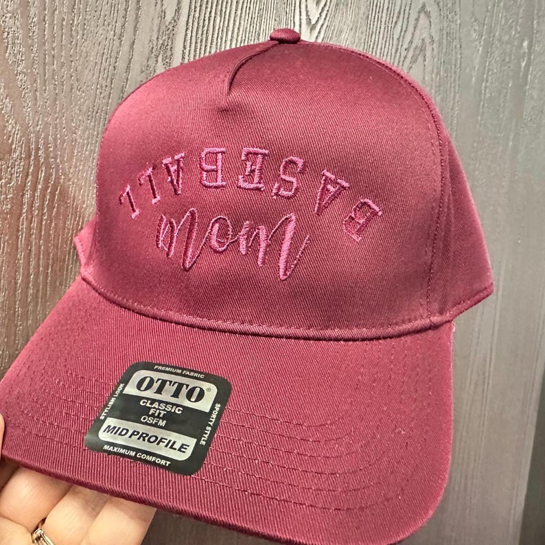 May include: A maroon baseball cap featuring the text BASEBALL mom embroidered in a lighter shade of maroon on the front. The cap includes a black sticker on the brim with the text OTTO CLASSIC FIT OSFM MID PROFILE MAXIMUM COMFORT.