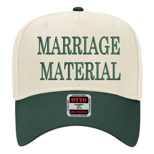 May include: A cream-colored baseball cap with a dark green brim. The words "MARRIAGE MATERIAL" are embroidered in dark green across the front. The cap has a curved bill and a button on top. The brand label "OTTO" is visible on the side.