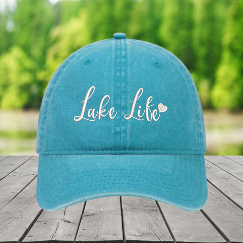 以下が含まれることがあります： ターコイズ色の野球帽。白い筆記体で「Lake Life」の文字が刺繍されています。「Life」の横には小さな白いハートがあります。帽子にはカーブしたつばと、上部にボタンが付いています。