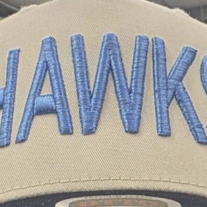 May include: A tan baseball cap featuring the word HAWKS embroidered in large, raised blue letters across the front crown. The cap has a structured design with visible stitching along the panels and a dark brim visible at the bottom edge.