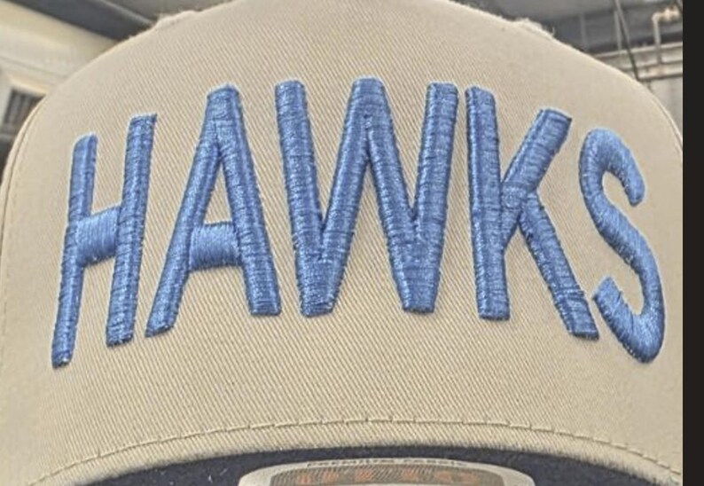 May include: A tan baseball cap featuring the word HAWKS embroidered in large, raised blue letters across the front crown. The cap has a structured design with visible stitching along the panels and a dark brim visible at the bottom edge.