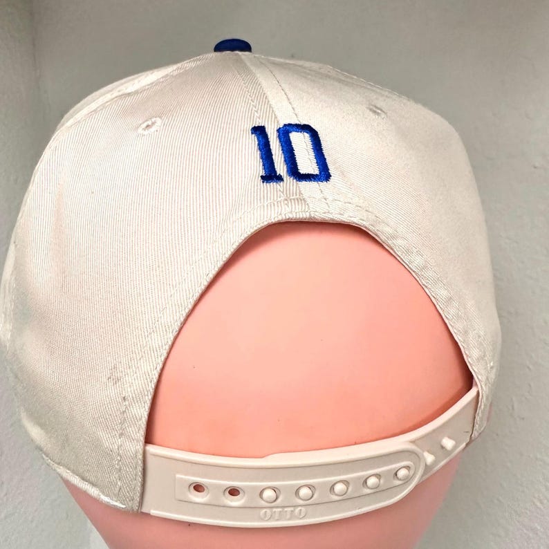 May include: White baseball cap with a blue number 10 embroidered on the back. The cap has a white adjustable strap with the word "OTTO" printed on it.