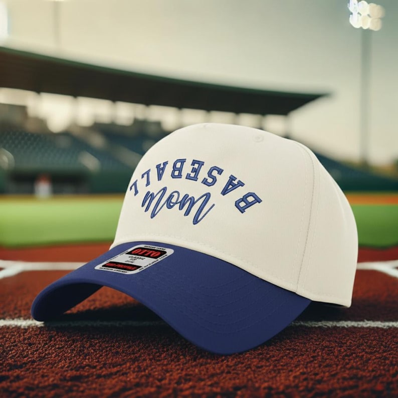 May include: A cream-colored baseball cap with a dark blue brim. The front of the cap features the text "BASEBALL" in a bold, arched blue font above the word "mom" in a blue cursive script. A small rectangular sticker is visible on the brim.
