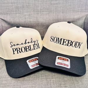 May include: Two baseball caps with a cream crown and a black brim. One cap says "Somebody's PROBLEM" in black script, and the other says "SOMEBODY". Both have an "OTTO" label.
