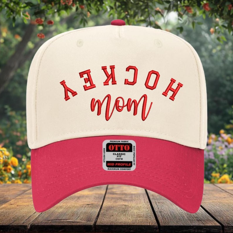 May include: A cream-colored baseball cap with a red brim and a red embroidered design on the front. The text reads HOCKEY in a block font and mom in a script font, both in red. A rectangular sticker on the brim displays the text OTTO.