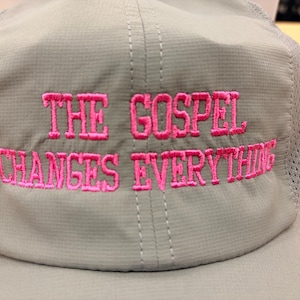 May include: A light gray baseball cap with mesh side panels and a structured front panel. The text THE GOSPEL CHANGES EVERYTHING is embroidered in bright pink thread across the front of the cap in a bold, capitalized, sans-serif font.