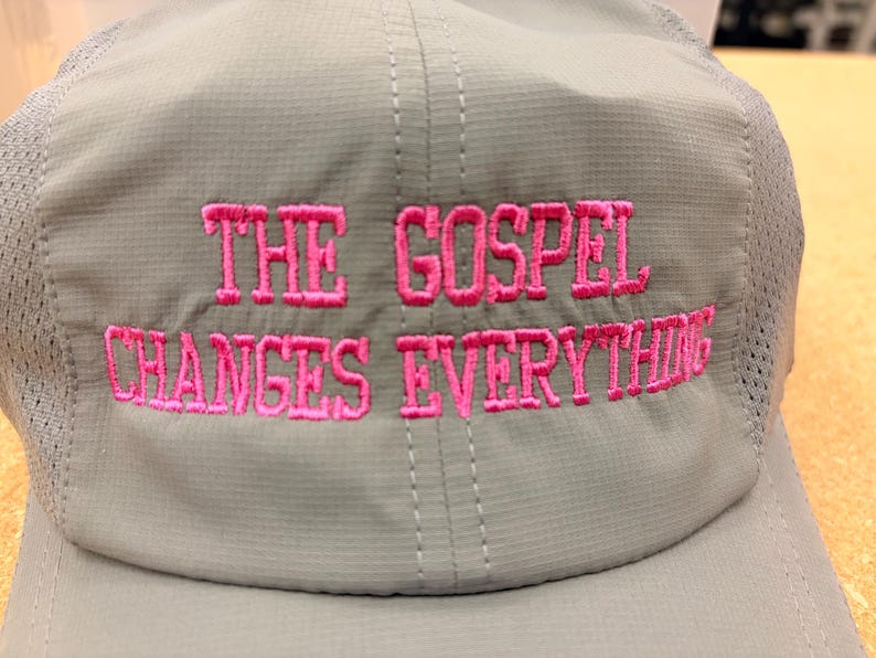 May include: A light gray baseball cap with mesh side panels and a structured front panel. The text THE GOSPEL CHANGES EVERYTHING is embroidered in bright pink thread across the front of the cap in a bold, capitalized, sans-serif font.