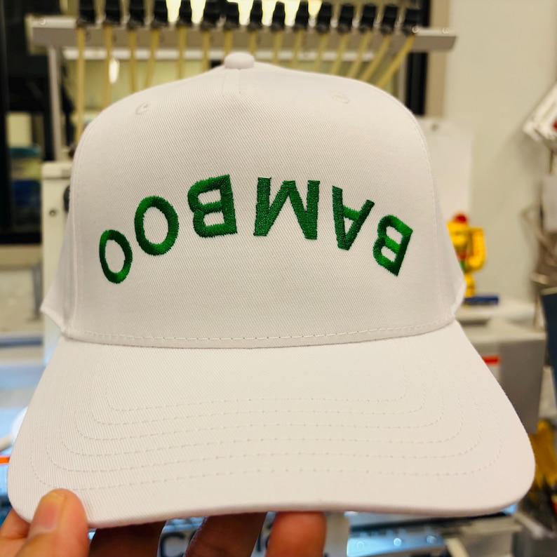 May include: A white baseball cap featuring the word BAMBOO embroidered in green thread across the front. The letters are oriented upside down, creating a distinct visual effect on the structured crown and the flat brim of the headwear.