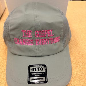 May include: A light gray five-panel cap featuring mesh side panels and the embroidered pink text THE GOSPEL CHANGES EVERYTHING on the front. A black rectangular sticker on the brim displays the brand name OTTO and the text PERFORMANCE OSFM RUNNING.