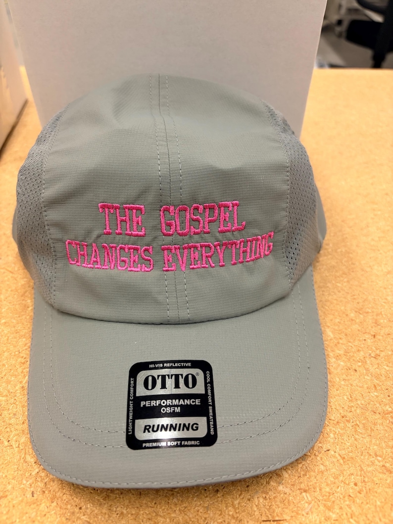 May include: A light gray five-panel cap featuring mesh side panels and the embroidered pink text THE GOSPEL CHANGES EVERYTHING on the front. A black rectangular sticker on the brim displays the brand name OTTO and the text PERFORMANCE OSFM RUNNING.