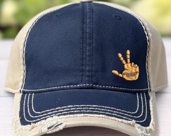 May include: A navy blue and beige baseball cap with a distressed, frayed edge on the brim. The front panel features an embroidered gold-colored hand making a peace sign, with a small vehicle grille design integrated into the palm.
