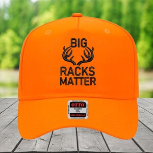 May include: Bright orange baseball cap with the text "BIG RACKS MATTER" in black. The cap features a curved brim and a structured crown. The cap has a tag that says "OTTO CLASSIC FIT MID PROFILE".