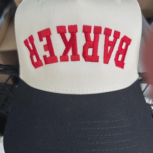 May include: A two-tone baseball cap featuring a cream-coloured front panel and a solid black brim. The front panel displays the word BARKER embroidered in bold, red, capital letters, positioned in an arched, upside-down orientation.