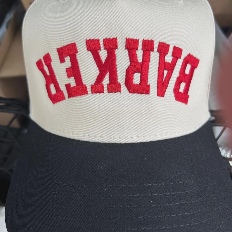 May include: A two-tone baseball cap featuring a cream-coloured front panel and a solid black brim. The front panel displays the word BARKER embroidered in bold, red, capital letters, positioned in an arched, upside-down orientation.