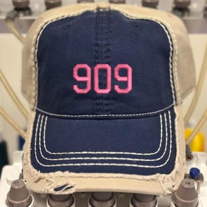 May include: A distressed baseball cap with a navy blue front panel and a tan mesh back. The number "909" is embroidered in pink on the front. White stitching outlines the brim and the cap's front panel. The cap has a pre-worn, vintage look.