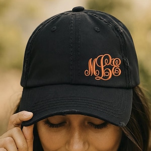 May include: Black baseball cap with an orange embroidered monogram. The cap has a distressed look and a curved brim. The monogram features elegant, swirling letters. A stylish accessory for casual wear.
